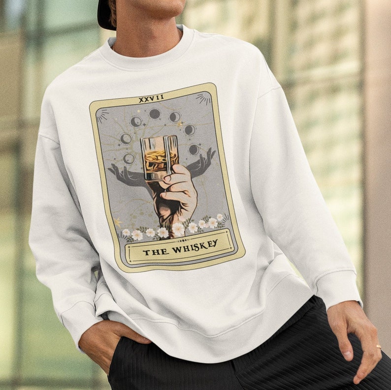 Może przedstawiać: Biała bluza z nadrukiem karty tarota. Karta nosi tytuł "The Whiskey" i przedstawia dłoń trzymającą szklankę whisky. Karta jest otoczona białą ramką i ma jasnoszary tło.