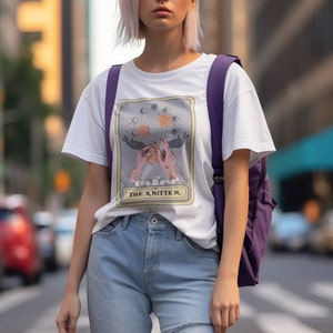 May include: White t-shirt with a graphic of a tarot card depicting two hands knitting with the text "The Knitter" below.