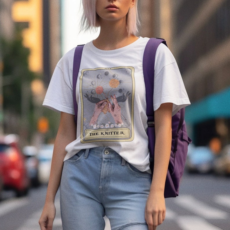 May include: White t-shirt with a graphic of a tarot card depicting two hands knitting with the text "The Knitter" below.