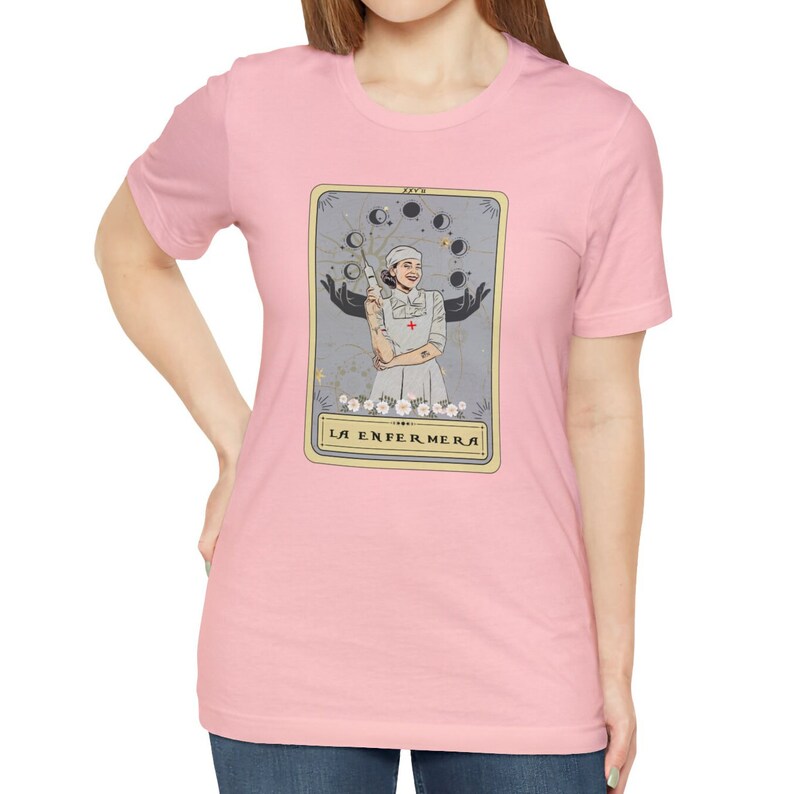 May include: A pink t-shirt featuring a tarot card design with a woman in a nurse's uniform holding a syringe. The card is titled "La Enfermera" in a gold frame.