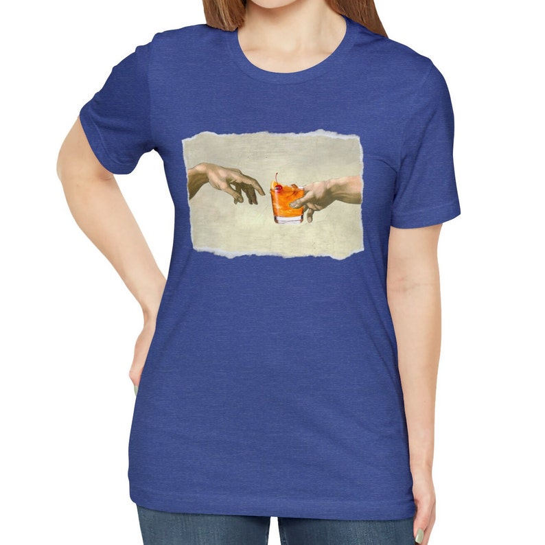 May include: A blue t-shirt with a white, torn-edged rectangle featuring a painting of two hands reaching towards each other, holding a glass of orange liquid with a cherry on top.