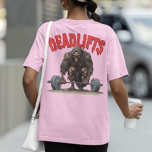 K&ouml;nnte beinhalten: Pinkes T-Shirt mit einem Grafikdruck eines Skeletts, das eine Langhantel hebt. Der Text "DEADLIFTS" ist in Rot &uuml;ber dem Skelett gedruckt.