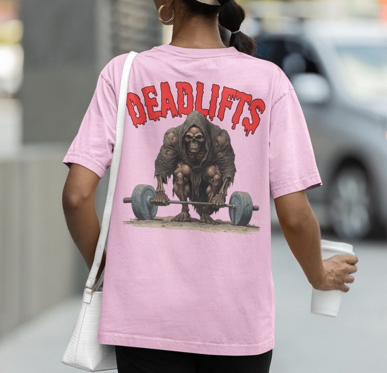 K&ouml;nnte beinhalten: Pinkes T-Shirt mit einem Grafikdruck eines Skeletts, das eine Langhantel hebt. Der Text "DEADLIFTS" ist in Rot &uuml;ber dem Skelett gedruckt.
