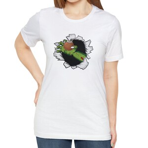 May include: White t-shirt with a graphic of a zombie hand reaching out of a ripped hole in the fabric, holding a green gourd.