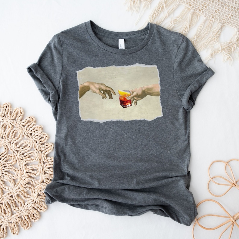 May include: A dark gray t-shirt with a print of two hands reaching towards each other, holding a glass of red liquid with a lemon wedge. The print is inspired by the famous Renaissance painting "The Creation of Adam".