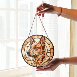 May include: Round stained glass panel featuring a dog's portrait. The dog has a red and white coat, with a black nose and eyes. The panel is held by a chain and has a black border. The background is a window with white curtains.