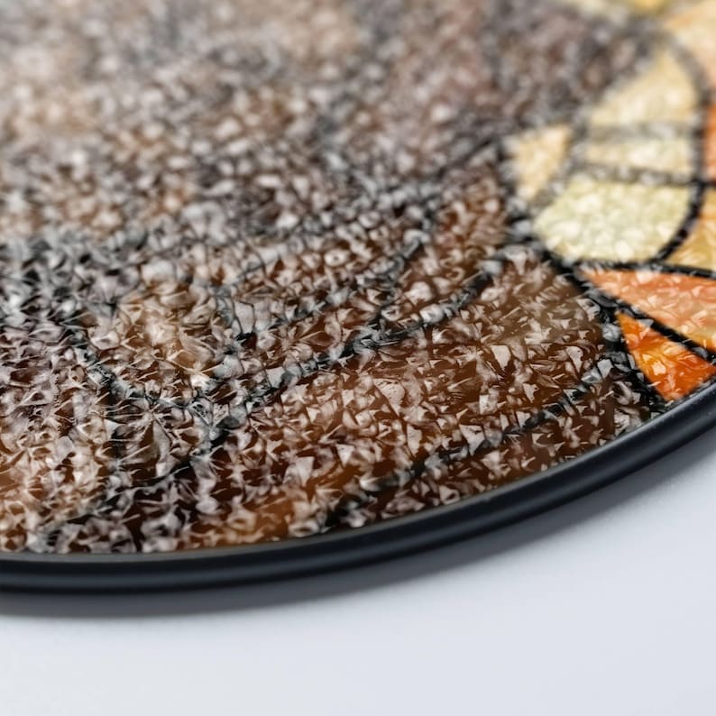 May include: Close-up of a decorative, round coaster with a textured, mosaic-like surface. The coaster features shades of brown, black, orange, and yellow, with a black border. The design has a unique, artistic appearance.