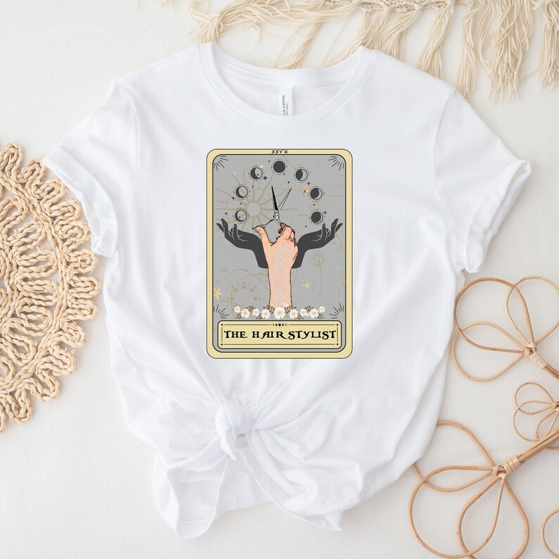 Puede incluir: Camiseta blanca con un gr&aacute;fico de una carta de tarot que muestra una mano sosteniendo tijeras y el texto "The Hair Stylist".
