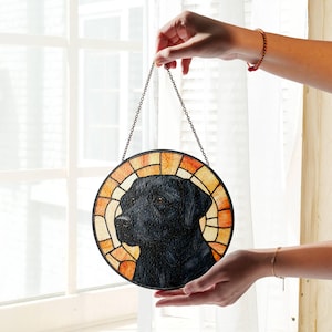 May include: Round stained glass window art featuring a black Labrador Retriever. The dog's portrait is centered within a circular design with a stained glass effect, using shades of orange, yellow, and brown. A metal chain is attached for hanging.
