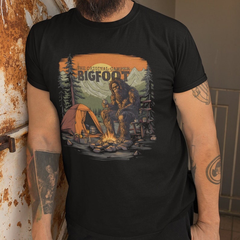 K&ouml;nnte beinhalten: Ein schwarzes T-Shirt mit einem Aufdruck von Bigfoot, der am Lagerfeuer im Wald sitzt. Der Text "The Original Camper Bigfoot" ist auf dem Shirt gedruckt.