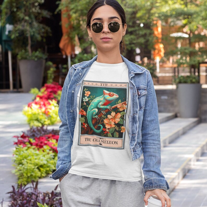 May include: White t-shirt with a graphic of a blue and green chameleon on a branch with pink and orange flowers. The graphic is surrounded by a vintage-style frame with the text "XVIII THE CHAMELEON".
