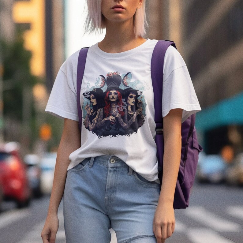 Puede incluir: Camiseta blanca con un estampado gr&aacute;fico de tres brujas con una luna creciente y estrellas. Las brujas llevan sombreros y t&uacute;nicas negras y sostienen varios objetos, entre ellos una caldera, una varita y un libro.