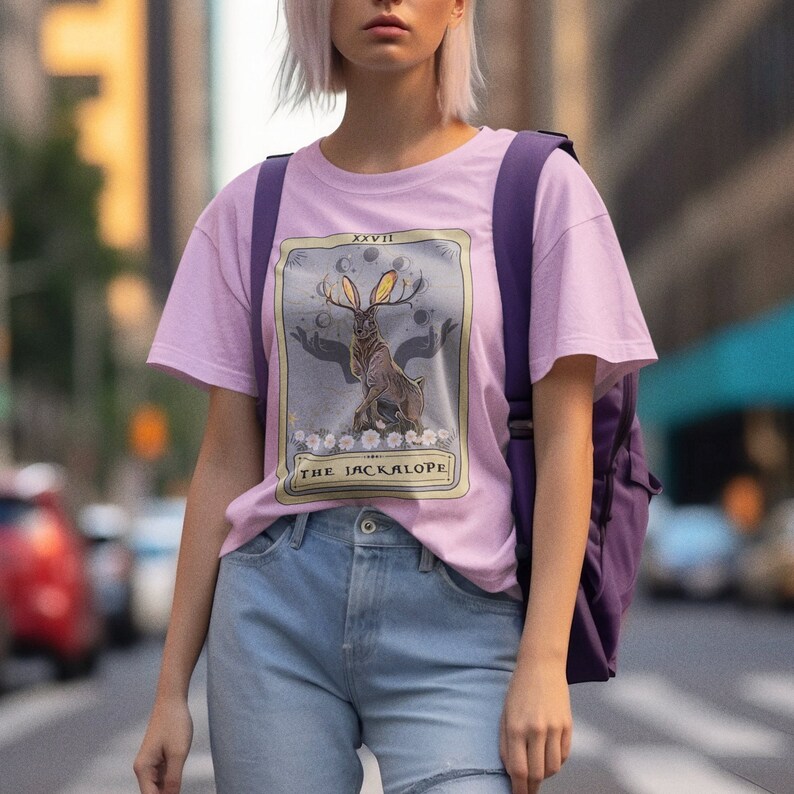 May include: A pink t-shirt with a graphic of a tarot card featuring a jackalope, a mythical rabbit with antlers, with the text "XXVII THE JACKALOPE".
