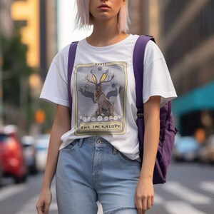 May include: White t-shirt with a graphic of a tarot card featuring a jackalope with antlers and the text "XXVIII THE JACKALOPE".