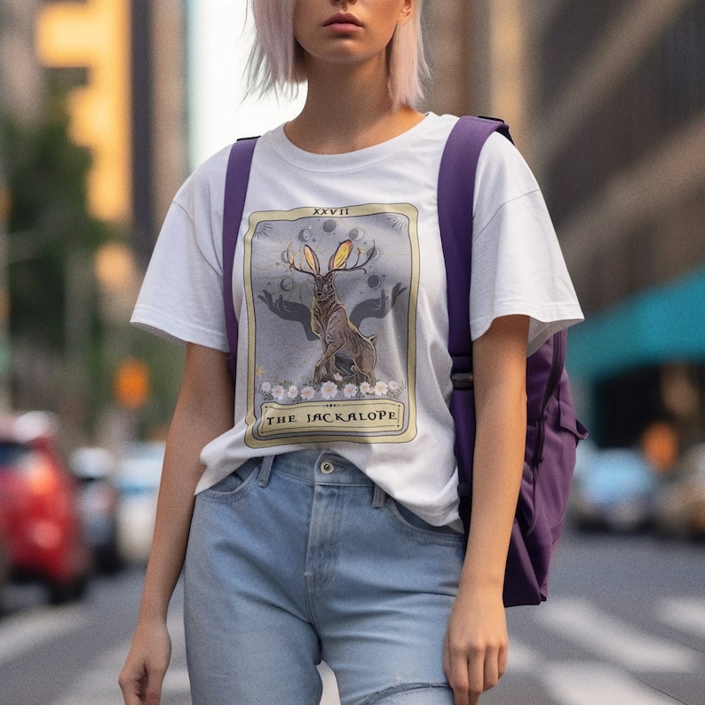 May include: White t-shirt with a graphic of a tarot card featuring a jackalope with antlers and the text "XXVIII THE JACKALOPE".