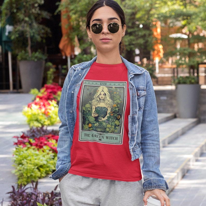 May include: A red t-shirt featuring a tarot card design with the text "THE GREEN WITCH." The design depicts a woman surrounded by greenery. The person is wearing a denim jacket and gray sweatpants.