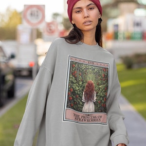 K&ouml;nnte beinhalten: Ein graues Sweatshirt mit einem grafischen Druck einer Tarotkarte. Die Karte tr&auml;gt den Titel "Die Prinzessin der Erdbeeren" und zeigt eine Frau mit roten Haaren, die durch einen Weg aus Erdbeerpflanzen geht.