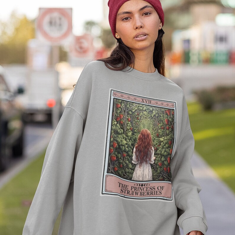 K&ouml;nnte beinhalten: Ein graues Sweatshirt mit einem grafischen Druck einer Tarotkarte. Die Karte tr&auml;gt den Titel "Die Prinzessin der Erdbeeren" und zeigt eine Frau mit roten Haaren, die durch einen Weg aus Erdbeerpflanzen geht.