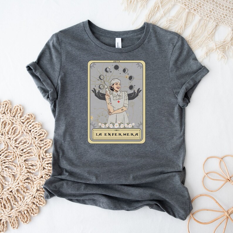 May include: A dark grey t-shirt with a tarot card design featuring a woman in a nurse's uniform holding a syringe. The card is titled "La Enfermera" in a gold frame.