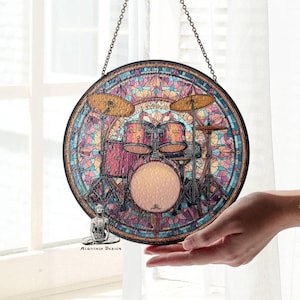 以下が含まれることがあります： ドラムセットを描いた丸いステンドグラスのアート作品。ピンク、青、金色のモザイク風のデザインです。チェーンで吊り下げられており、「Alquimia Design」の文字が入っています。