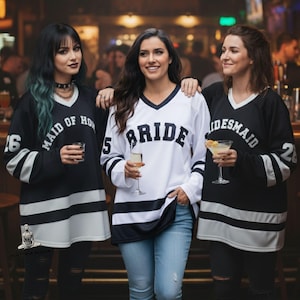 Pu&ograve; includere: Tre persone indossano maglie in stile hockey bianche e nere. La persona al centro indossa una maglia bianca con la scritta "BRIDE" sul davanti. Le altre due indossano maglie nere con "MAID OF HONOR" e "BRIDESMAID". Ogni maglia ha un numero sulla manica.