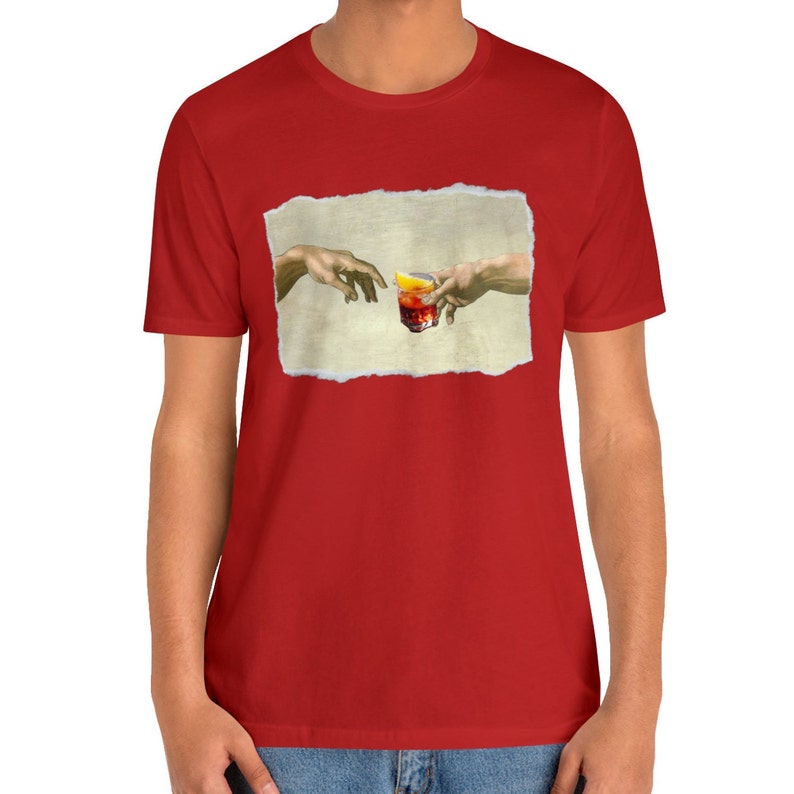 May include: A red t-shirt with a white, torn-paper frame featuring a painting of two hands reaching towards each other, holding a glass of orange liquid with a lemon wedge.