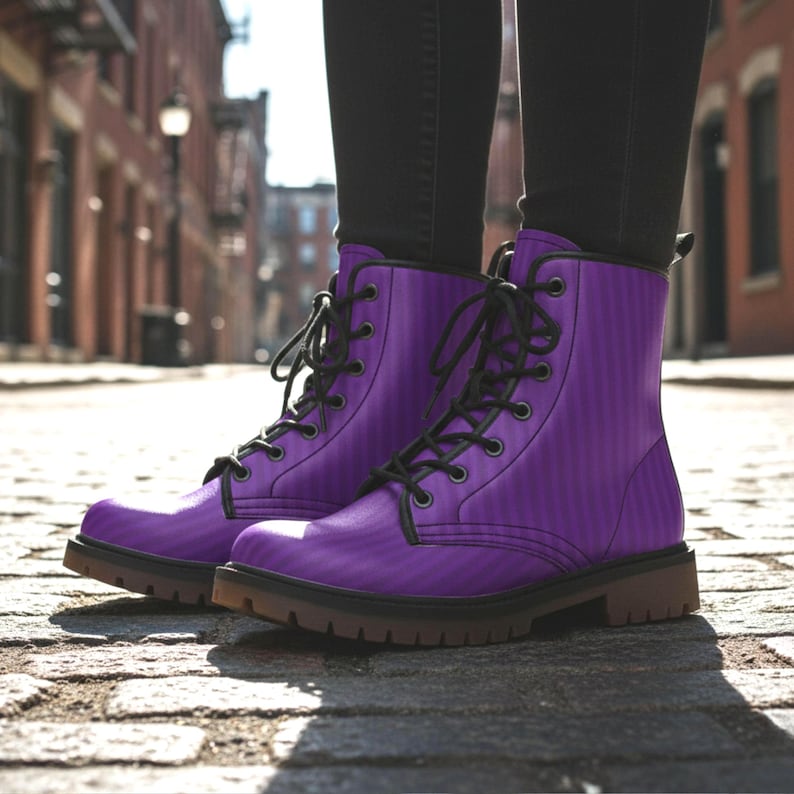 May include: A pair of purple combat boots with black laces. The boots have a vertical striped pattern and a brown sole. The boots are worn with black pants and are on a cobblestone street.