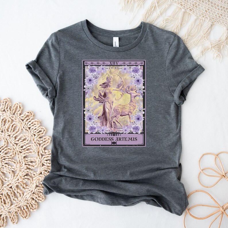 Może przedstawiać: Szary t-shirt z nadrukiem przedstawiającym fioletową i ż&oacute;łtą ilustrację bogini z łukiem i strzałami oraz jeleniem. Ilustracja jest otoczona fioletowymi kwiatami, a także tekstem "XIV", "GODDESS ARTEMIS" i symbolem p&oacute;łksiężyca.