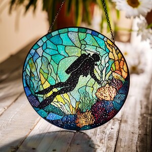 Op de afbeelding: Een gebrandschilderd raam dat aan een ketting hangt, met een silhouet van een duiker die door een koraalrif zwemt. Het glas is een mix van blauw, groen, geel en oranje.