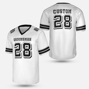 May include: White football jersey with black accents, featuring the word "GROOMSMAN" and the number "28" on the front. The back of the jersey displays "CUSTOM" and the number "28". The jersey has black stripes on the sleeves.