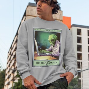 Peut inclure: Un sweat-shirt gris avec un imprim&eacute; graphique d'un extraterrestre portant un sweat &agrave; capuche et tapant sur un ordinateur portable. Le graphique est dans le style d'une carte de tarot avec le texte "The Programmer" et "XVIII" en haut.