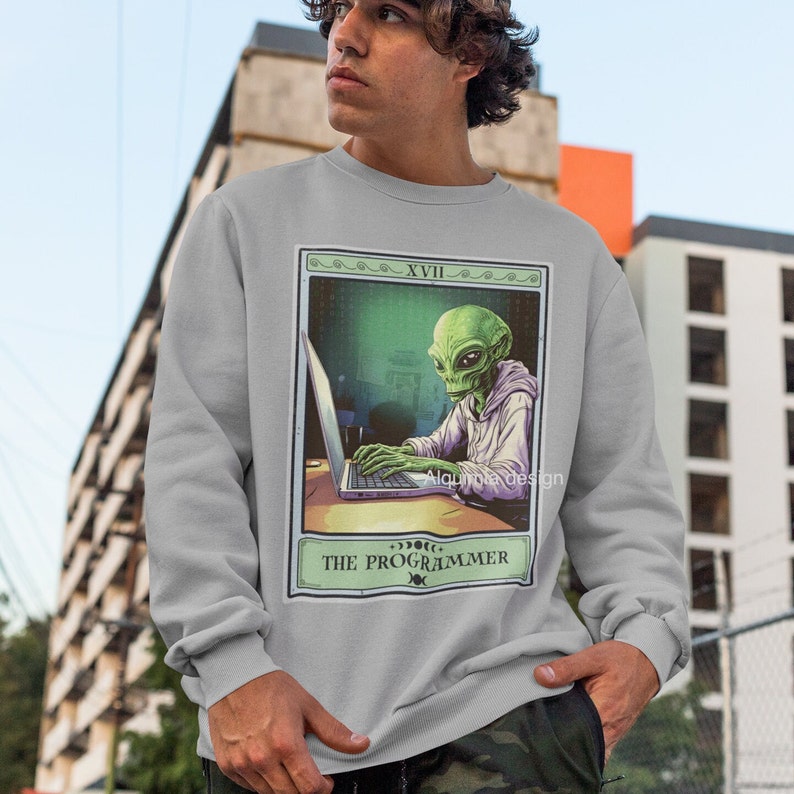 Peut inclure: Un sweat-shirt gris avec un imprim&eacute; graphique d'un extraterrestre portant un sweat &agrave; capuche et tapant sur un ordinateur portable. Le graphique est dans le style d'une carte de tarot avec le texte "The Programmer" et "XVIII" en haut.