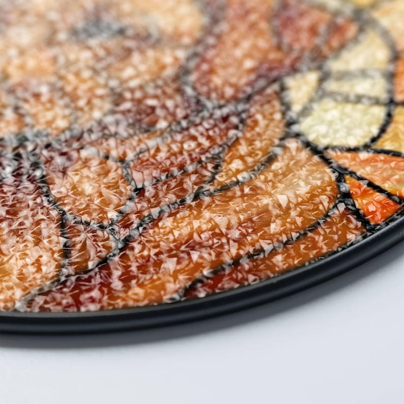 May include: Close-up of a decorative plate with a mosaic design. The plate features a black rim and a textured surface with shades of orange, brown, and yellow. The mosaic pattern is outlined with dark lines, creating a stained-glass effect.