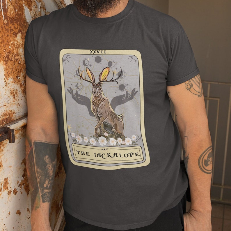 May include: A dark gray t-shirt with a vintage tarot card design featuring a jackalope with antlers and a crescent moon. The card is labeled "XXVII THE JACKALOPE".