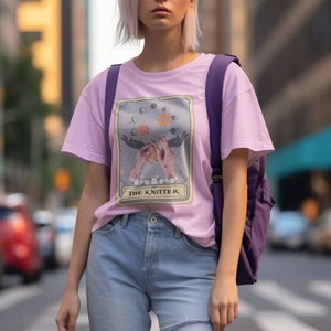 May include: A pink t-shirt with a graphic of a tarot card featuring a pair of hands knitting with a moon and star design. The card reads "The Knitter".