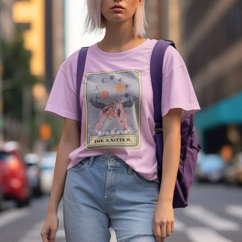 May include: A pink t-shirt with a graphic of a tarot card featuring a pair of hands knitting with a moon and star design. The card reads "The Knitter".
