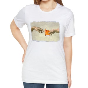 May include: A white t-shirt with a graphic of two hands reaching towards each other, holding a glass of orange liquid with a cherry on top. The graphic is inspired by the famous Renaissance painting "The Creation of Adam".