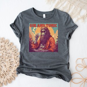 May include: A dark gray t-shirt with a graphic of a cartoon Bigfoot holding a drink and smoking a joint. The text "Gin and Tonic" is printed above the image.