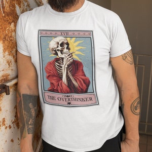 May include: White t-shirt with a tarot card design featuring a skeleton in a red robe, smoking a cigarette. The card is labeled "XVII The Overthinker".