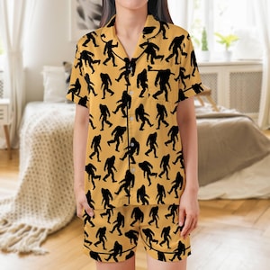 Puede incluir: Conjunto de pijama amarillo mostaza con un patrón de silueta negra repetida de Bigfoot. El conjunto incluye una camisa con botones de manga corta y pantalones cortos a juego. El pijama tiene ribete negro.