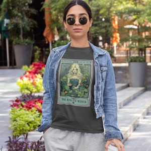 May include: Dark gray t-shirt featuring a tarot card design with a blonde woman surrounded by greenery. The text "THE GREEN WITCH" is below the image. The shirt is worn with a denim jacket and gray sweatpants.