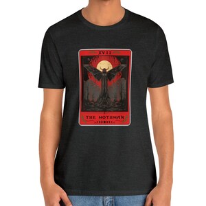 May include: A dark heather grey t-shirt featuring a red and black tarot card design. The card depicts a black mothman with large wings, a red moon, and the text "XVII THE MOTHMAN".