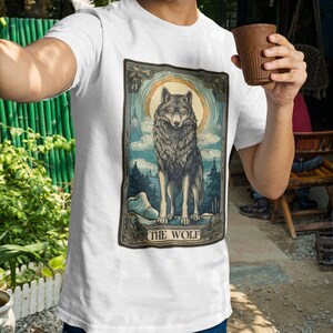 May include: White t-shirt with a graphic print of a wolf standing in front of a full moon. The graphic is surrounded by a decorative frame and the text "THE WOLF" is printed below the wolf.