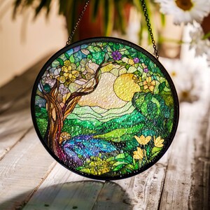 Op de afbeelding: Een hangend glas-in-loodraam met een rond ontwerp met een boom, bloemen en een zon in een groen en geel landschap.