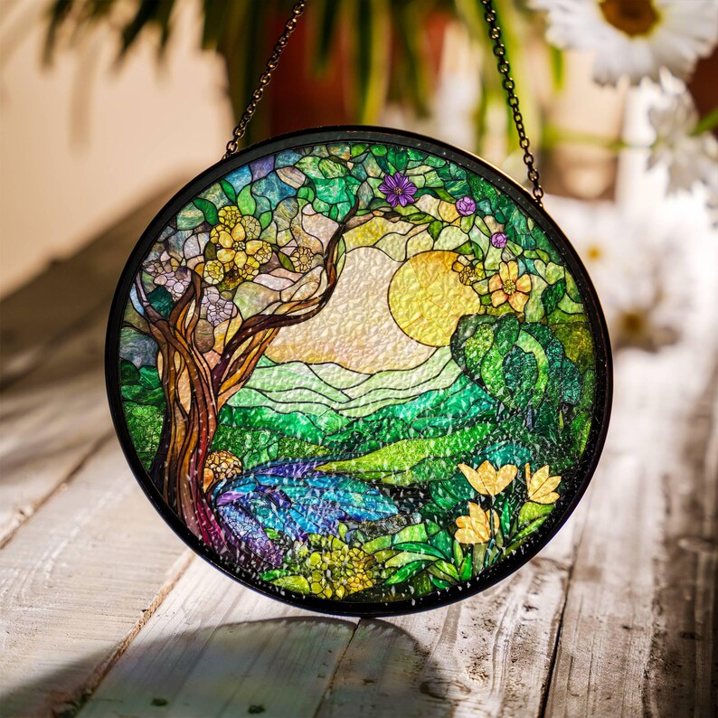 Op de afbeelding: Een hangend glas-in-loodraam met een rond ontwerp met een boom, bloemen en een zon in een groen en geel landschap.