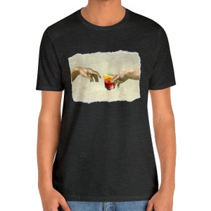 May include: A dark heather grey t-shirt with a graphic of two hands reaching towards each other, one holding a glass of red liquid with a lemon wedge. The graphic is inspired by the famous Renaissance painting "The Creation of Adam" by Michelangelo.