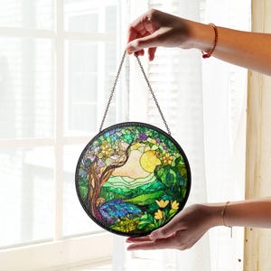 Op de afbeelding: Een rond glas-in-loodraam dat aan een ketting hangt. Het glas-in-lood toont een boom, een zon en een landschap met groene heuvels en gele bloemen.