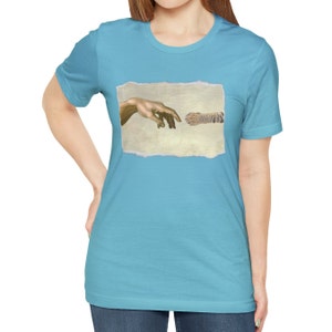 May include: A light blue t-shirt with a graphic of a hand reaching out to touch another hand, referencing the famous "The Creation of Adam" painting. The graphic is in a distressed, ripped paper style.