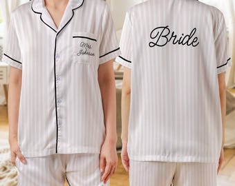 May include: A white and pink striped pajama set. The short-sleeved top has a pocket with "Mrs. Johnson" embroidered on it. The shorts have "Bride" embroidered on the leg. The back of the top says "Bride". The set has black trim.