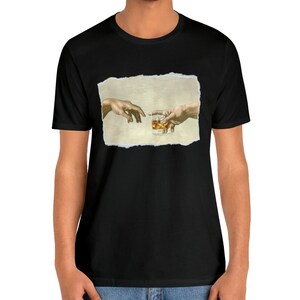 K&ouml;nnte beinhalten: Ein schwarzes T-Shirt mit einem Aufdruck von zwei H&auml;nden, die sich gegenseitig erreichen, wobei eine ein Glas Whisky h&auml;lt. Der Druck befindet sich auf einem strukturierten, beigefarbenen Hintergrund.